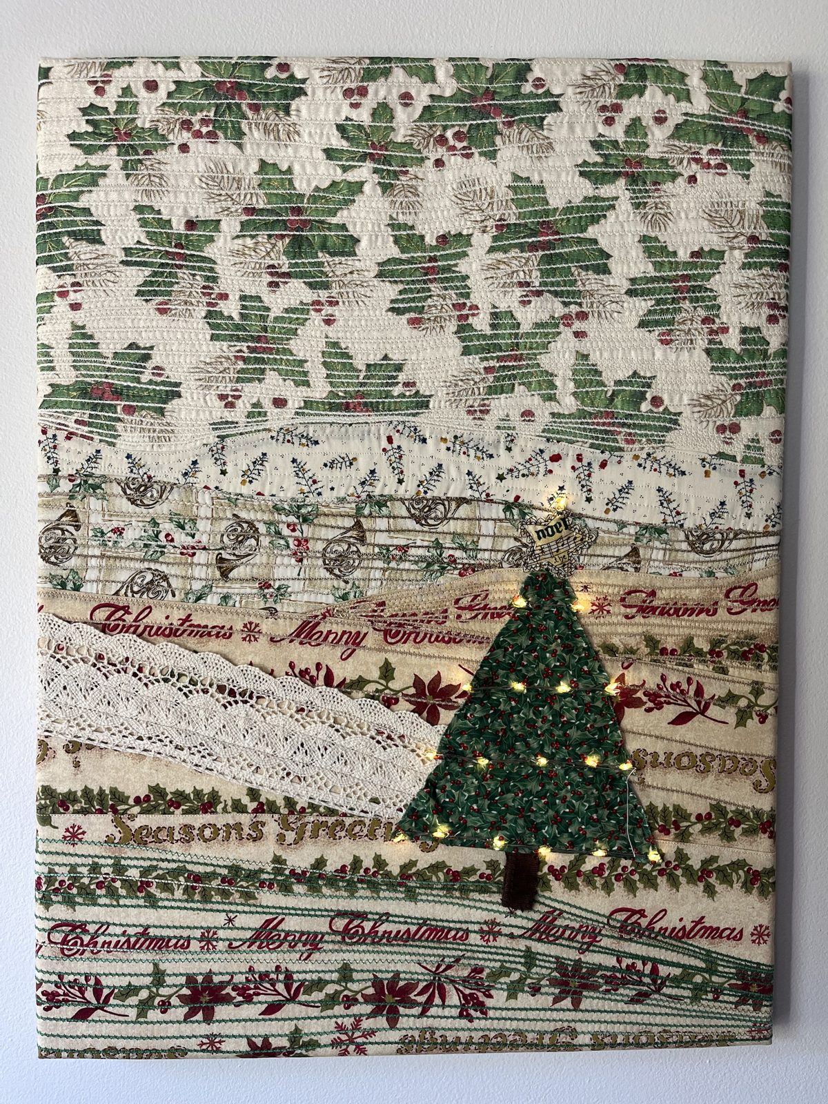 Vertical Christmas quilt: layered holiday-print ridges, vintage lace, and a green fabric tree with tiny fairy lights and a Noel star