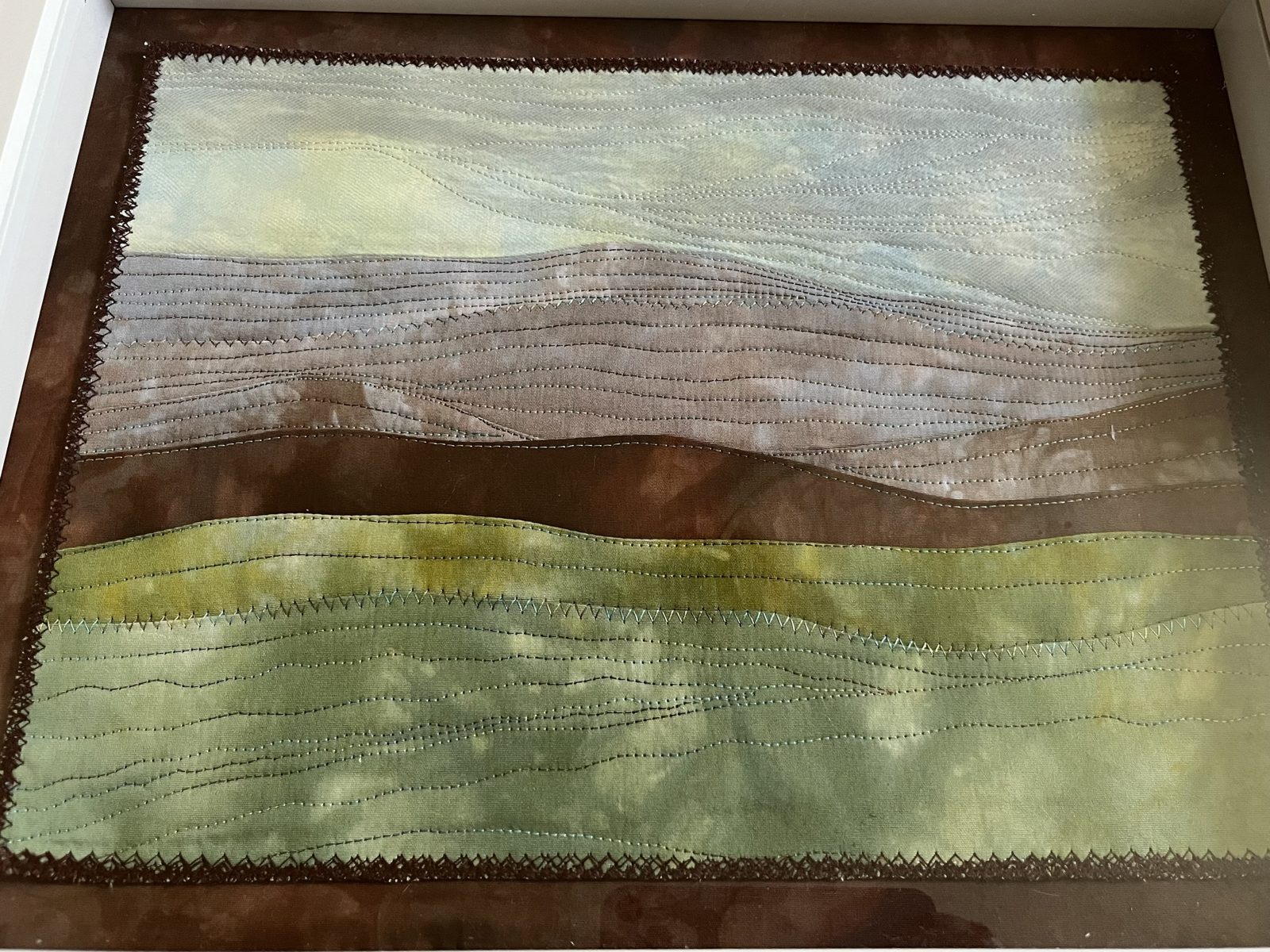 Framed landscape quilt in hand-dyed greens, mauves and browns, covered in wavy lines of quilting with a zigzag edge