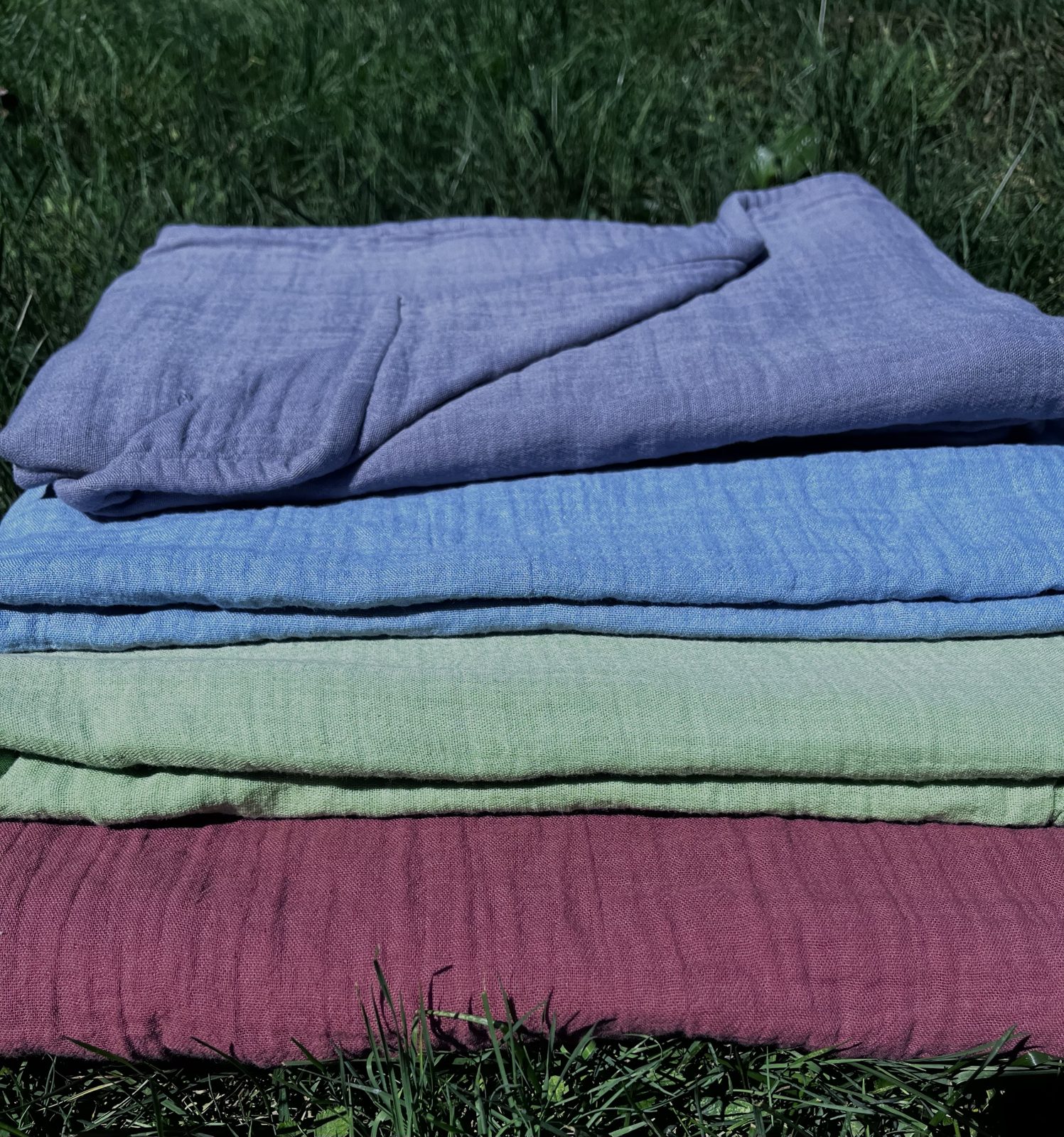 Stack of hand-dyed double gauze in periwinkle, cornflower blue, sage green and plum, on the grass
