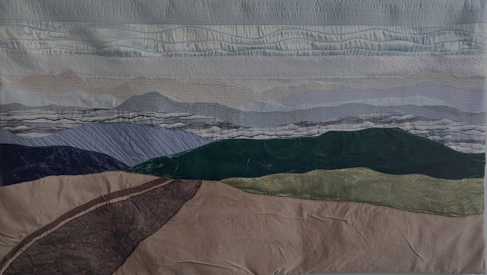 Wide landscape quilt: pale quilted sky, a band of sequin mesh, layered blue-grey ridges, a dark green velvet mountain and a winding road in the foreground
